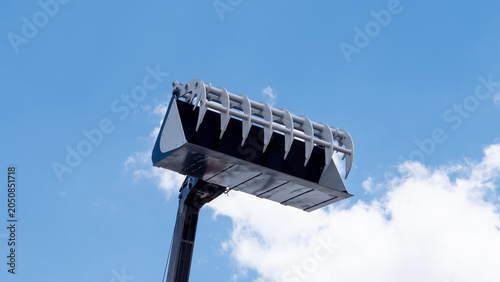 A large excavator bucket is lifted high against a clear blue sky with soft white clouds, showing the metal teeth and industrial equipment from a low angle
