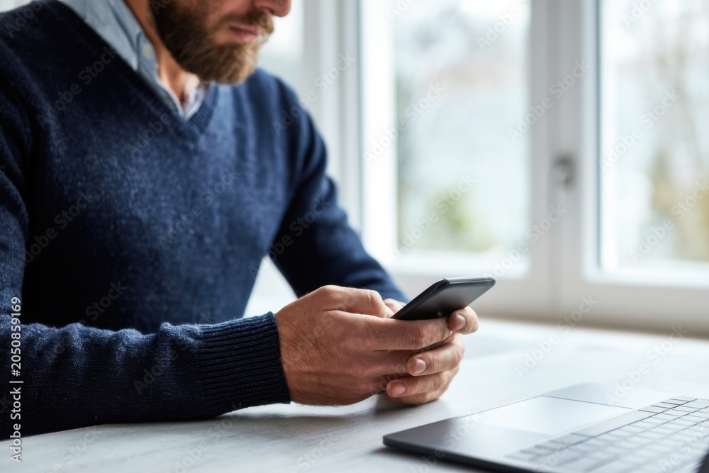 Fototapeta premium The Smartphone in Hands of Bearded Man at Bright Home Office Desk