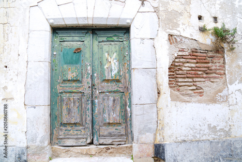 An old weathered green wooden door with peeling paint set in a stone archway on a distressed white plaster wall showing exposed red bricks in the historic town of Elvas, Alentejo, Portugal.