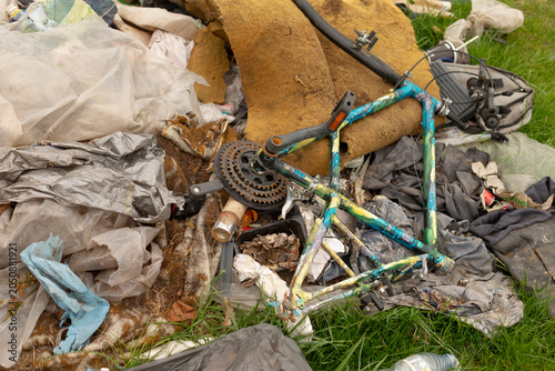 Discarded bicycle frame with corroded chainring and sprockets lying among torn fabric, plastic bags, crushed cans and upholstery foam on patchy grass under