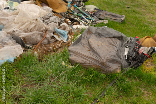 A pile of discarded household waste scattered on a grassy field including torn plastic bags, fabric scraps, broken bicycle parts and mixed debris. The scene