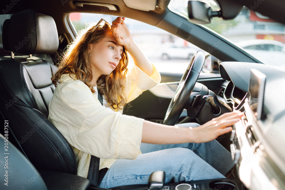 Fototapeta premium A young woman sits behind the wheel of a hot car. A female driver feels unwell due to the heat in the car on a sunny day. Concept of air conditioning, cooling, and transportation.