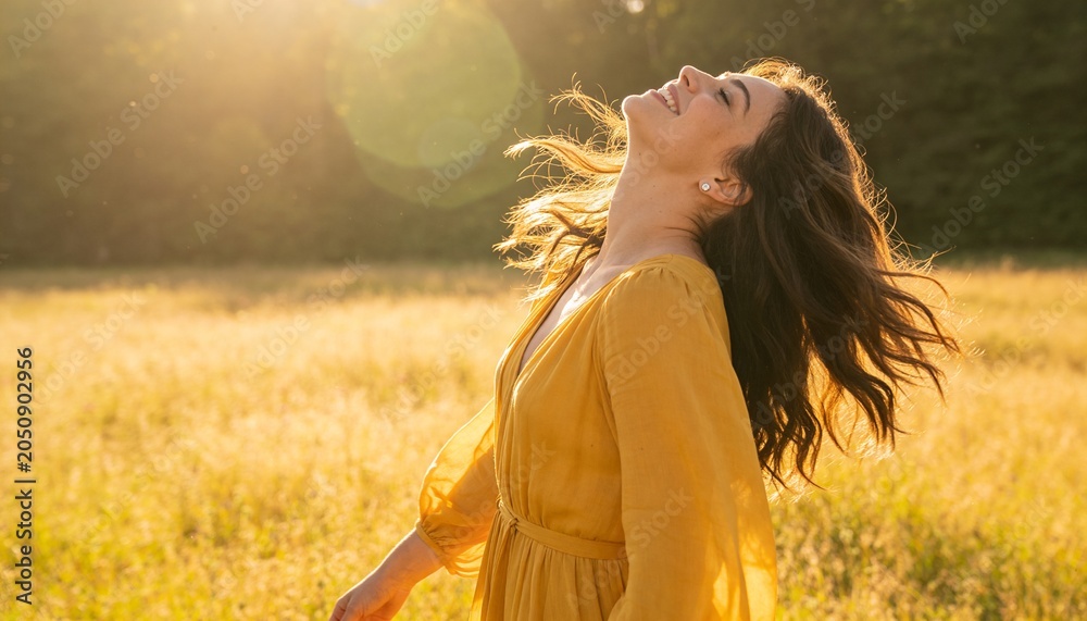 Obraz premium Tilting woman in mustard dress and studs in golden meadow amid tall grasses, catching lens flare