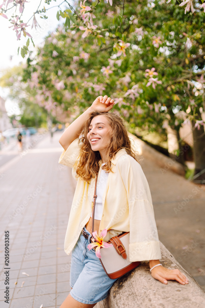 Naklejka premium A smiling woman enjoys a sunny day in a park, surrounded by blooming trees. The woman, in casual clothes, poses outdoors. Concept of beauty, fashion, and relaxation.