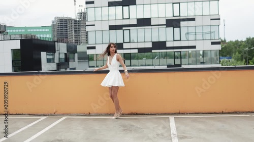 Stylish young woman in white mini dress standing on rooftop parking lot with modern city buildings background, turning around to face camera. Urban fashion lifestyle concept for apparel, beauty and ci
