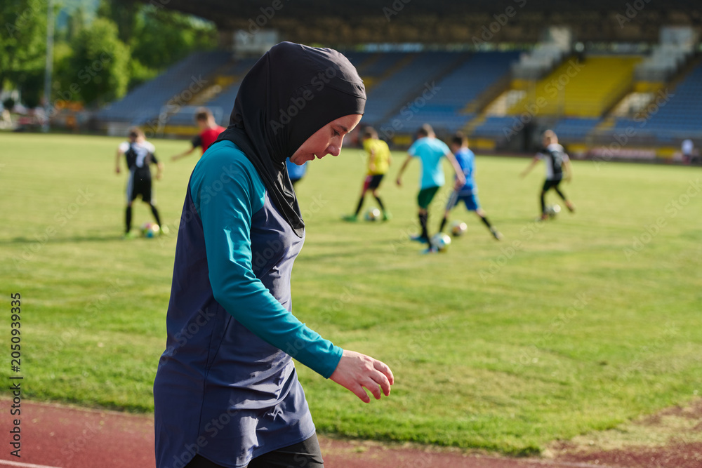 Obraz premium Muslim Woman Coach Wearing Hijab Walking Along Soccer Field While Youth Team Trains