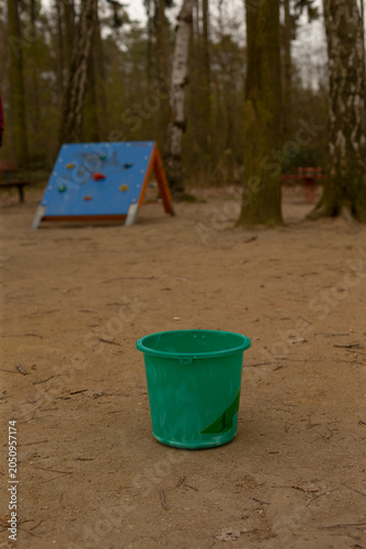 A single green plastic bucket sits on a sandy playground surface in a wooded park. In the midground there is a small children's climbing wall painted blue