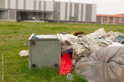 A pile of discarded household items and rubbish scattered on a grassy lot in an urban area. The foreground shows a small white appliance tipped on its side