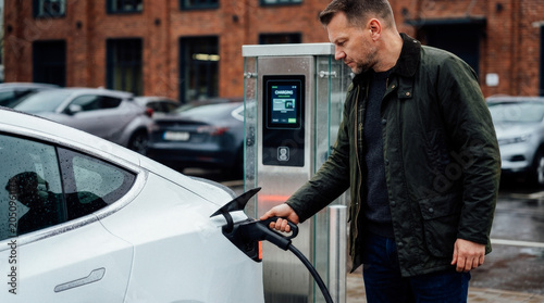 Man charging electric car at charging station, focusing on sustainable transportation and modern technology in an urban parking lot.