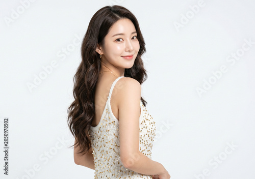 A beautiful young Asian model with long wavy black hair smiles warmly as she looks back over her shoulder while wearing an elegant white lace strap dress against a plain white background