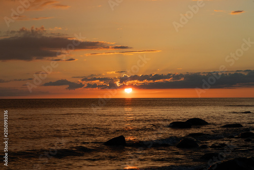 Sunset Over Rocky Shoreline With Golden Sky, Calm Ocean Reflections And Dramatic Clouds