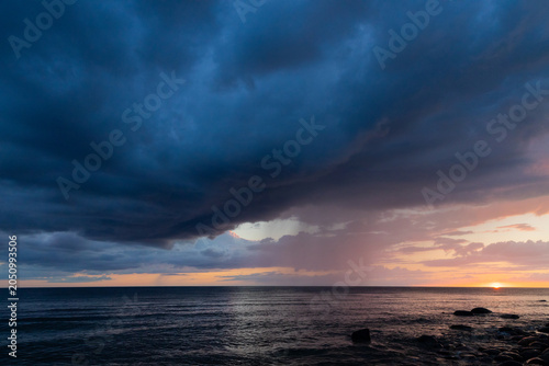 Dramatic Storm Over Ocean At Sunset With Rocky Shoreline And Rainfall On Horizon