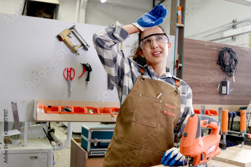 Exhausted woman carpenter with safety goggles and an apron wiping sweat off forehead during making wooden furniture. Tired beautiful female craft worker working in woodworking carpentry workshop.