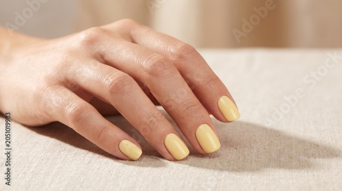 A close-up of a woman's hand resting on a light-colored surface. The nails are painted in a soft yellow shade. The skin appears smooth and well-groomed.