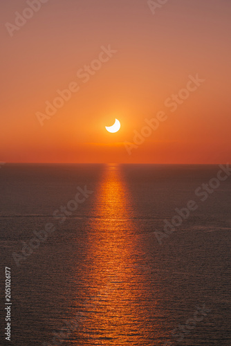 Partial solar eclipse at sunset over calm ocean with bright sun path reflection on water