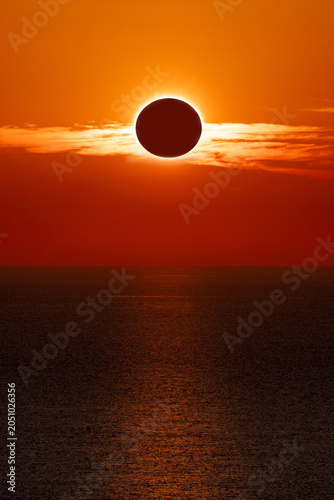 Total solar eclipse at sunset over the ocean, with a spectacular orange sky and the sun's reflection on the water.