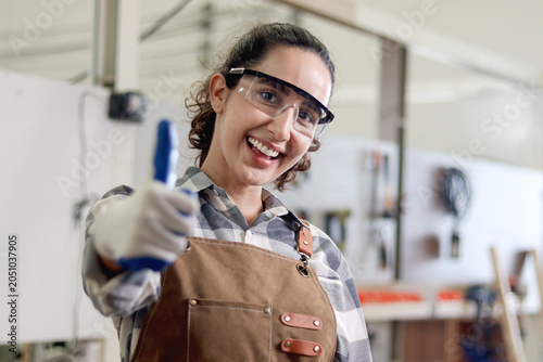 Photos Portrait of happy smiling woman carpenter wears safety glasses goggles and apron, giving thump up, standing at carpentry workshop