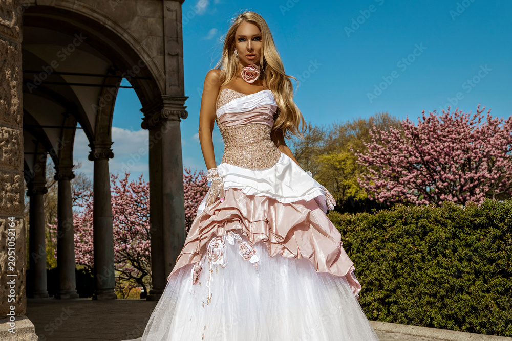 Fototapeta premium Stunning blonde fashion model in an extravagant white and pink ballgown posing outdoors under a bright blue sky with blooming spring blossoms and stone arches.