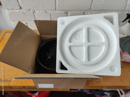 Open cardboard box with black cooking pot and white foam packaging on wooden table.