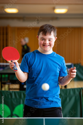 A young boy with Down syndrome enjoys a game of table tennis in a lively gym. His joyful expression shows his excitement as he prepares to play. Generative AI.
