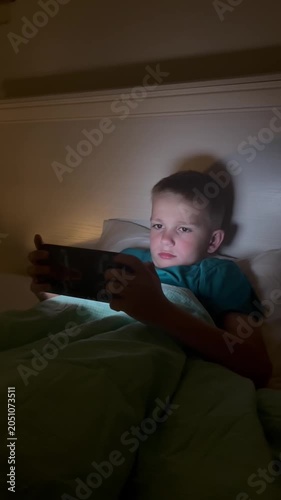 Vertical. A child holds a smartphone under the blanket's light, scrolling through content. The boy uses gadgets while getting ready for bed, his face illuminated by the blue light of the smartphone sc