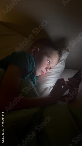 Vertical. A child holds a smartphone under the blanket's light, scrolling through content. The boy uses gadgets while getting ready for bed, his face illuminated by the blue light of the smartphone sc