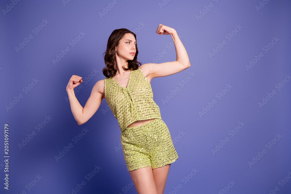 Fototapeta premium Young woman flexing her muscles in a green crochet outfit against a purple background, showing confidence, empowerment and playful fashion attitude