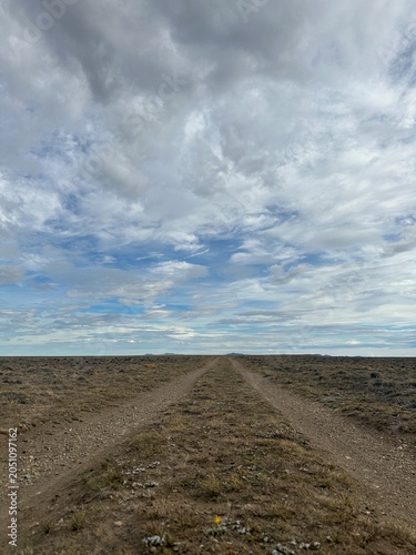 Remote dirt road crossing the vast Patagonian steppe in Argentina beneath a dramatic cloudy sky. A minimalist landscape that conveys solitude, freedom, adventure, and the immense scale of nature.