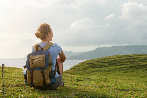 Back view of woman with canvas backpack sitting on green grass hill, contemplating mountains and sea. Solo travel concept.