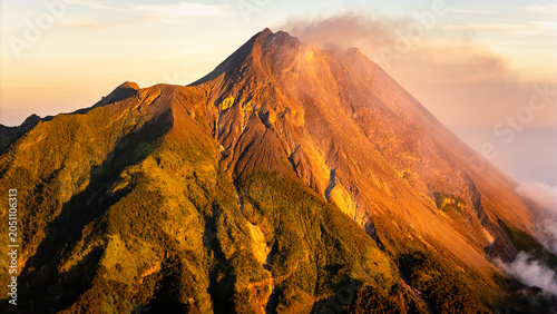 Aerial view of Mount Merapi volcano with smoke rising from the crater and golden sunlight illuminating the rocky slopes and green ridges in Indonesia.