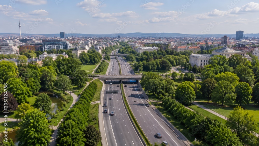 Fototapeta premium An elevated perspective captures a modern highway bustling with cars, flanked by lush green parks and urban architecture, with a prominent tower visible in the distance
