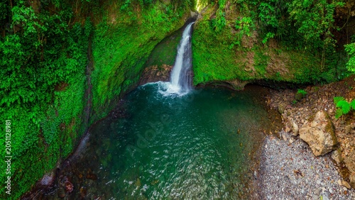 Aerial view of Palak Siring Waterfall cascading into a turquoise pool surrounded by lush green mossy cliffs in Kecamatan Arma Jaya, Bengkulu, Indonesia.