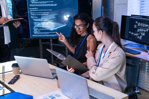 Focused female software engineer discuss project script via laptop. Diverse tech startup coder woman use tablet to check computer program code and web database development in office meeting
