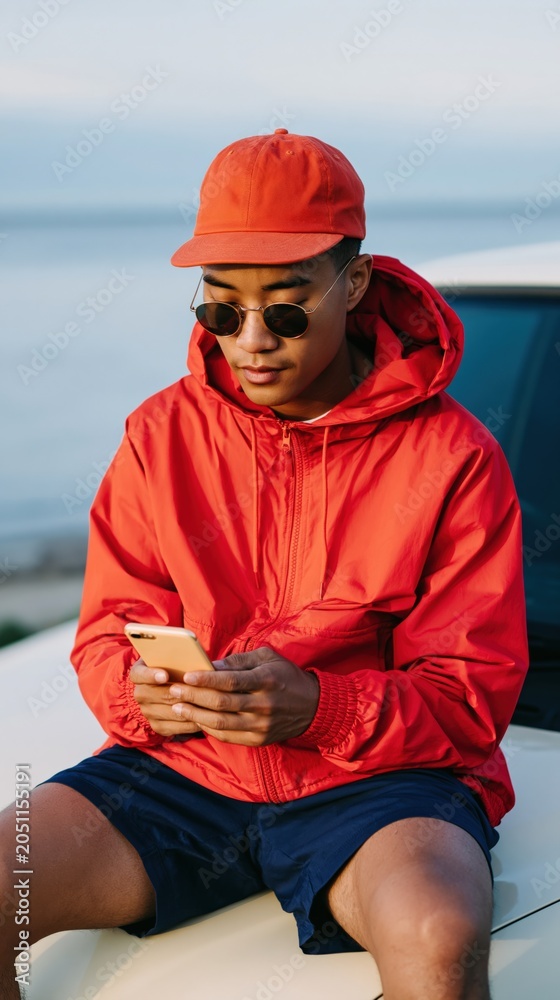 Obraz premium Casual young person in bright red jacket and cap using smartphone on car hood by the ocean for lifestyle banner and social media background