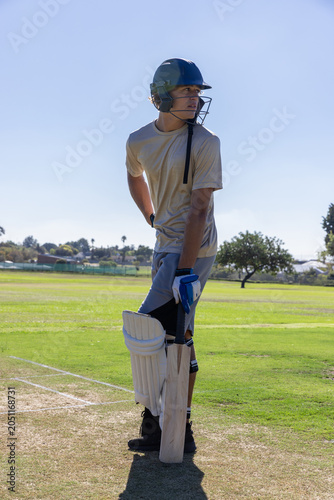 Cricket batsman is standing on marked pitch holding bat with pads and helmet under sunshine