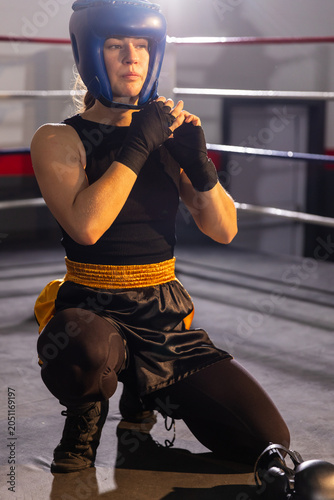 Boxer is kneeling and fastening blue headgear on canvas ring floor with gloves nearby