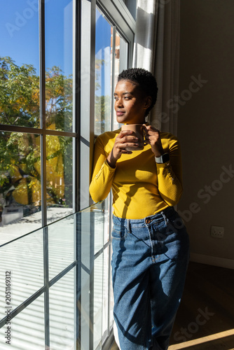 African American woman standing by multi-pane window at home holding ceramic mug in yellow top