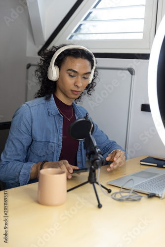 Non-binary creator wearing denim shirt and headset preparing mic in attic, holding phone