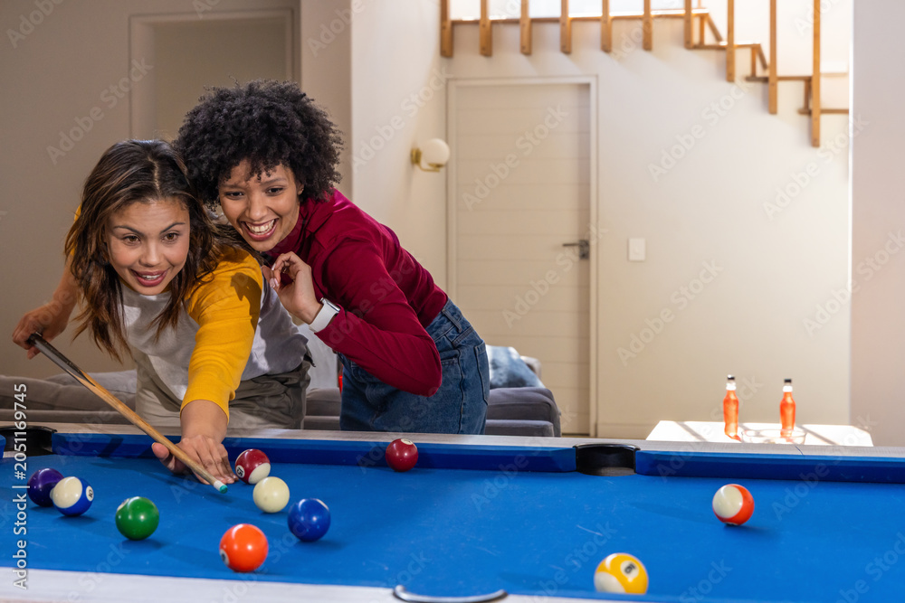 Fototapeta premium Diverse female friends holding cue stick, lining up shot on blue pool table in living room