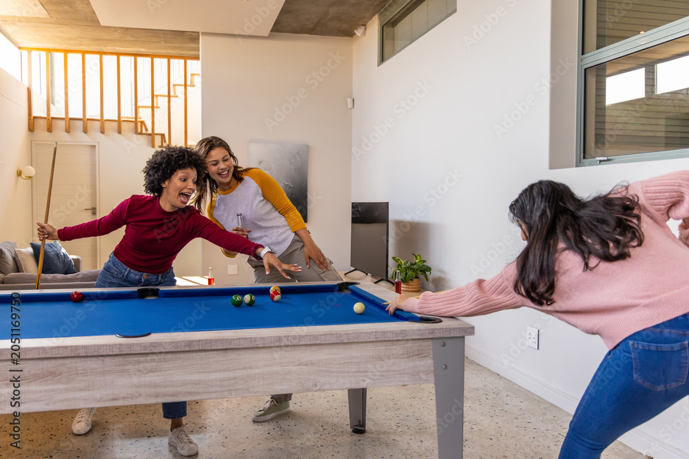 Obraz premium Three female friends playing pool on blue-felt pool table at home, using wooden cues now