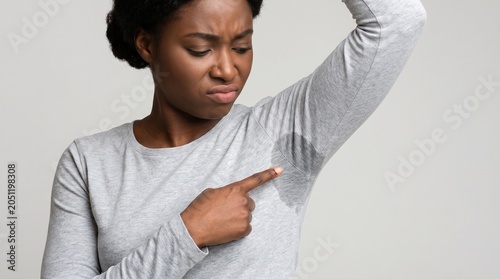 A young woman with dark skin looking concerned while pointing at a large sweat stain on her gray shirt sleeve