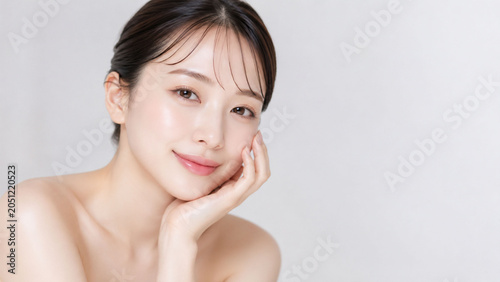 ライトグレーのミニマルな背景の前で、頬に手を添えてやさしく微笑む若い女性を捉えた、上品で清潔感のあるナチュラルビューティーポートレート。透明感のある素肌、やわらかな艶感、整えたまとめ髪、ヌーディーで洗練された表情が印象的で、スキンケア、保湿化粧品、美容液、素肌美、エステ、フェイスケア広告に適した商業向け人物素材