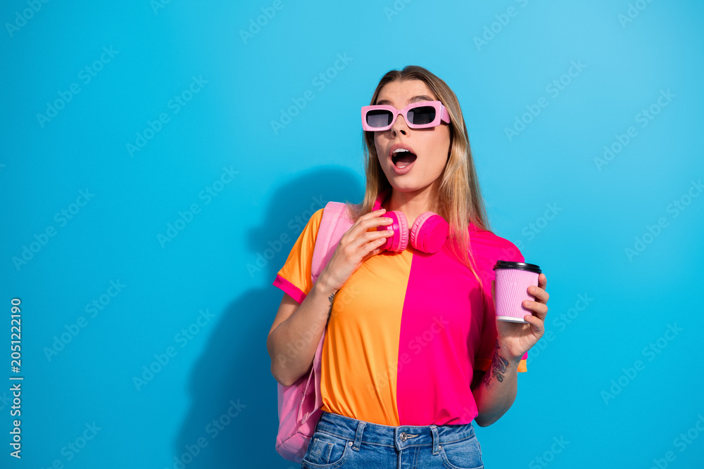 Fototapeta premium Young woman in colorful outfit with pink headphones holding coffee on blue background lifestyle model