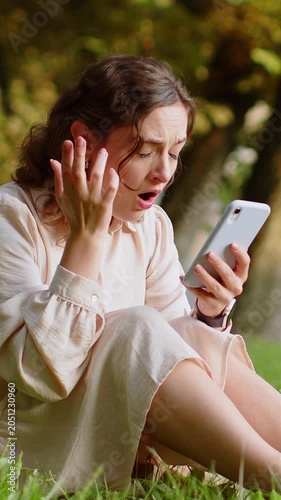 Young woman use smartphone typing browsing, loses becoming surprised sudden lottery results, bad news, fortune loss, fail. Lady sitting on grass in urban sunset city park outdoors. Town lifestyles