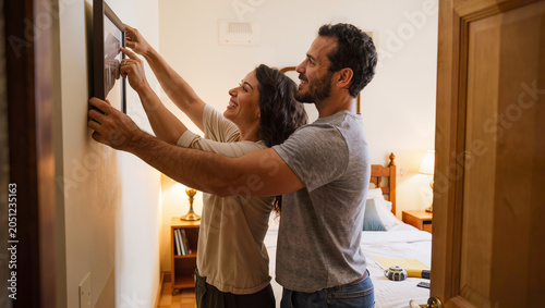 Brazilian wife hanging framed picture on bedroom wall with husband help in new home