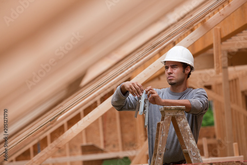 Hispanic carpenter standing on a ladder holding metal reinforcement bracing at a house under