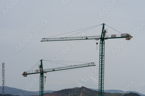 Two green tower cranes rise above a hilly landscape under an overcast sky, showing construction equipment, development, and changing urban edges.