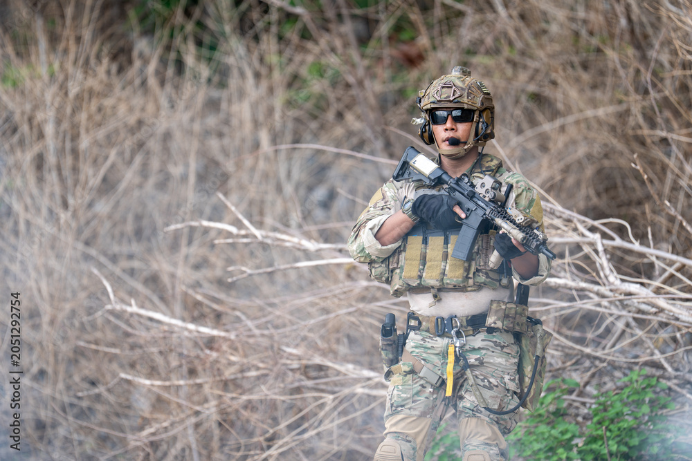 Fototapeta premium Soldier in camouflage uniform aiming rifle gun while kneeling near rock cliff. Tactical military warrior holding weapon during combat training outdoor