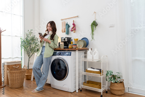 Young Asian woman using smartphone during laundry routine at home. Smiling female resting near automatic washing machine inside modern room houseworks lifestyle concept
