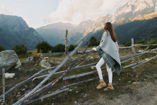 Schilderij op canvas Young woman with long hair wearing a gray shawl and white pants climbs over a wooden fence in a mountainous rural area during daytime
